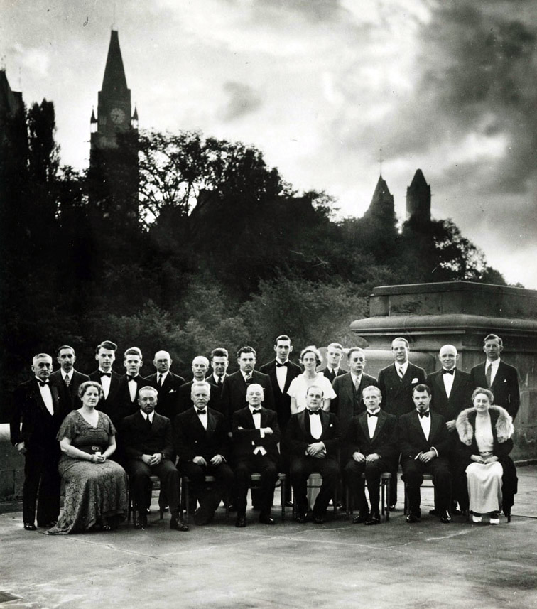 Black-and-white photograph of 20 men and 3 women, all wearing suits and dresses posing for the camera. In the background is a line of trees; the Canadian Houses of Parliament are partly visible, including the large carillon clock tower