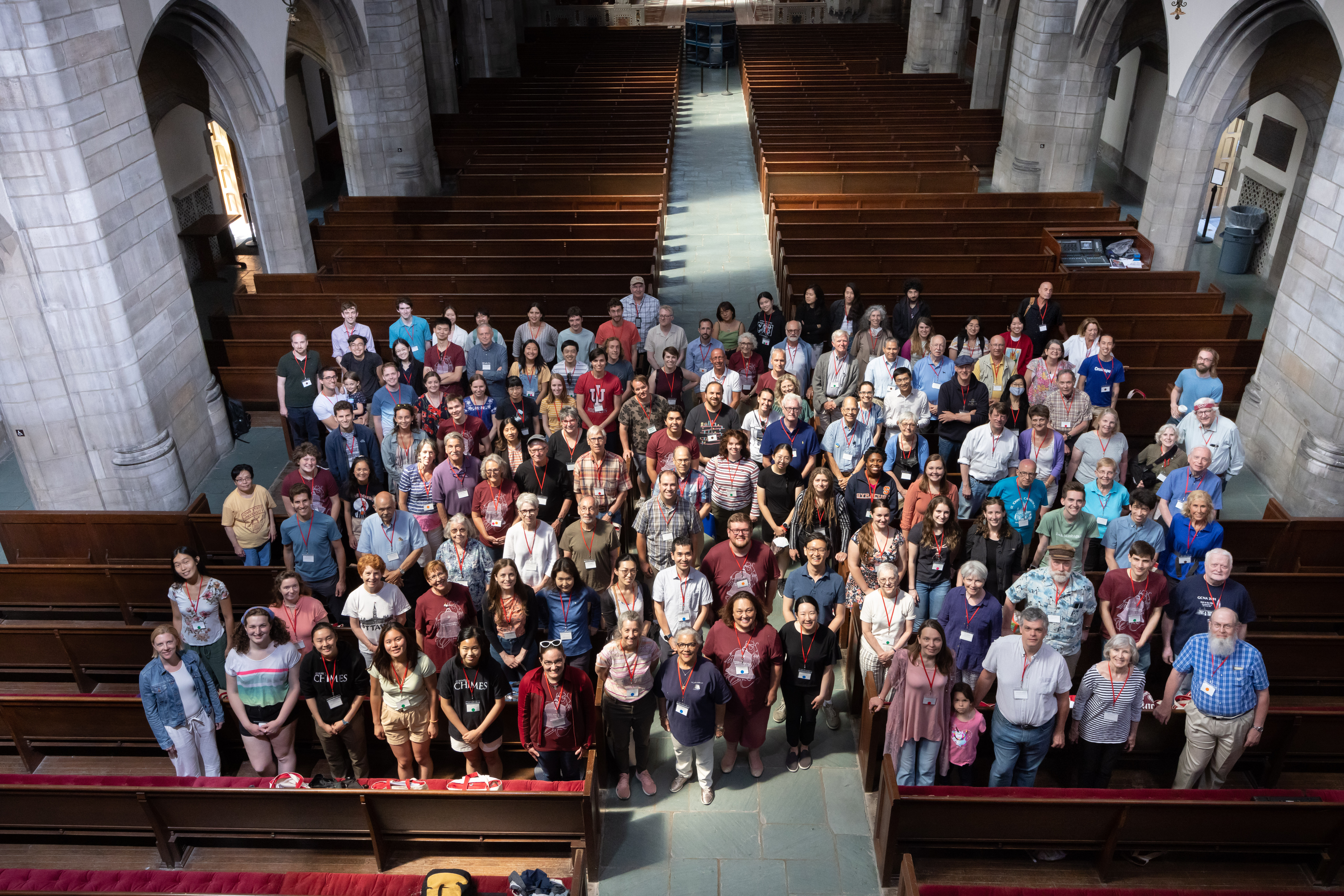 Group photograph of nearly 140 people in a large stone church standing between several wooden pews and smiling up at the camera